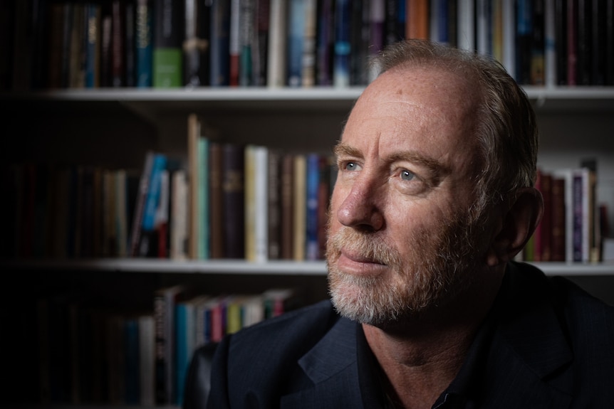 A man looks up, with a bookshelf behind him.