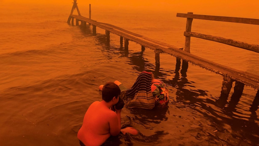 A boy hunkers down in seawater near what looks like an old pier, the sky an evil red from bushfire activity.