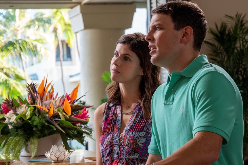 A young white couple look concerned and are standing in a lobby filled with palms and exotic flowers