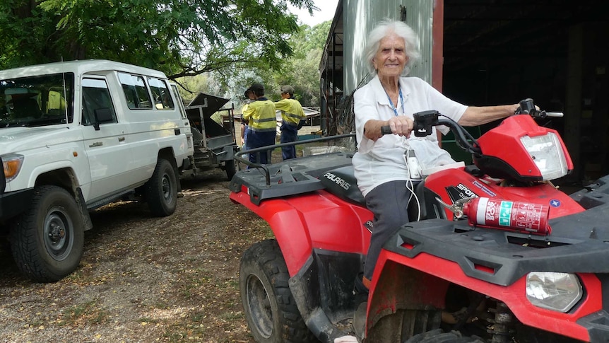 A woman sits on a quad bike outside a shed while men gather behind her.