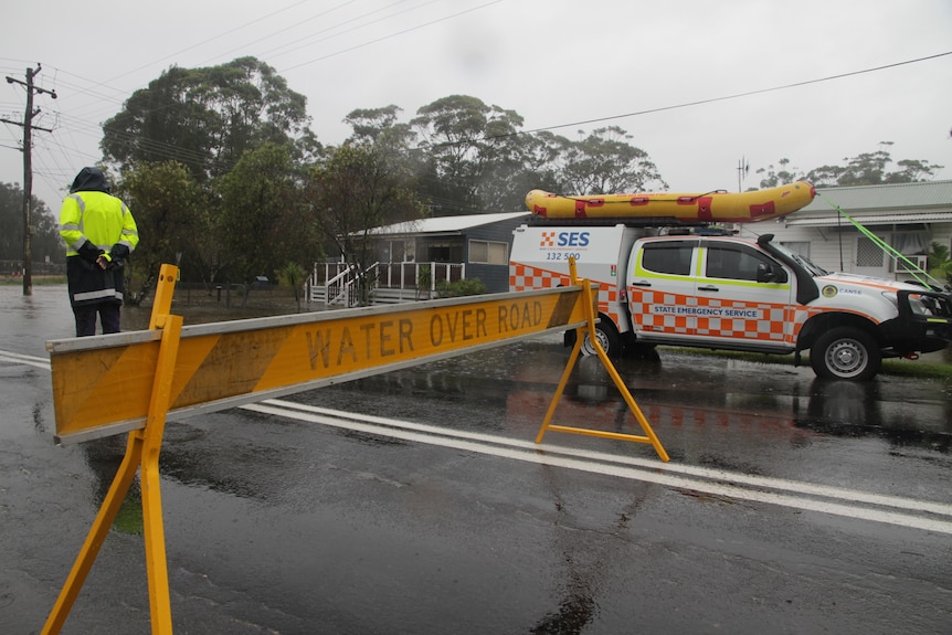 Man wearing yellow high-vis standing in front of 'water over road' sign, SES vehicle next to house, water seen on road