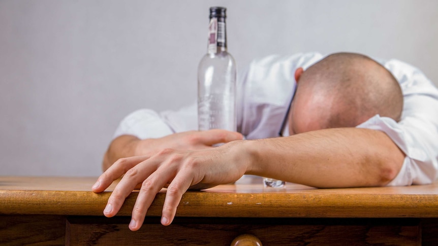 A man holding a bottle of alcohol is seen slumped over a desk.