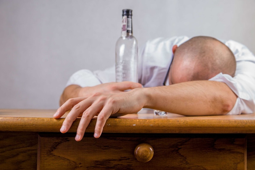 A man holding a bottle of alcohol is seen slumped over a desk.