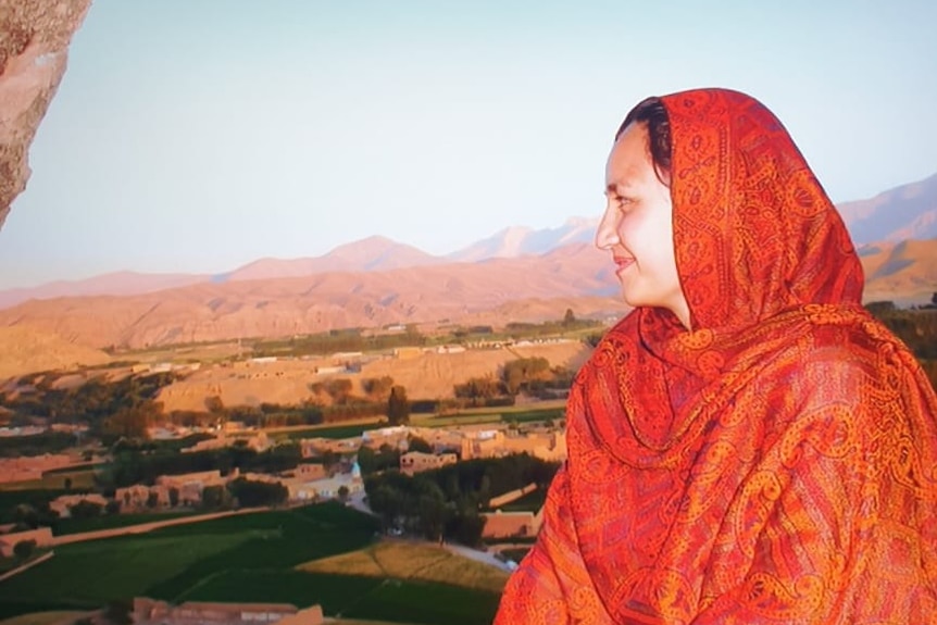 A woman wearing a brightly-coloured hijab looks over an Afghan landscape, with mountains in the distance