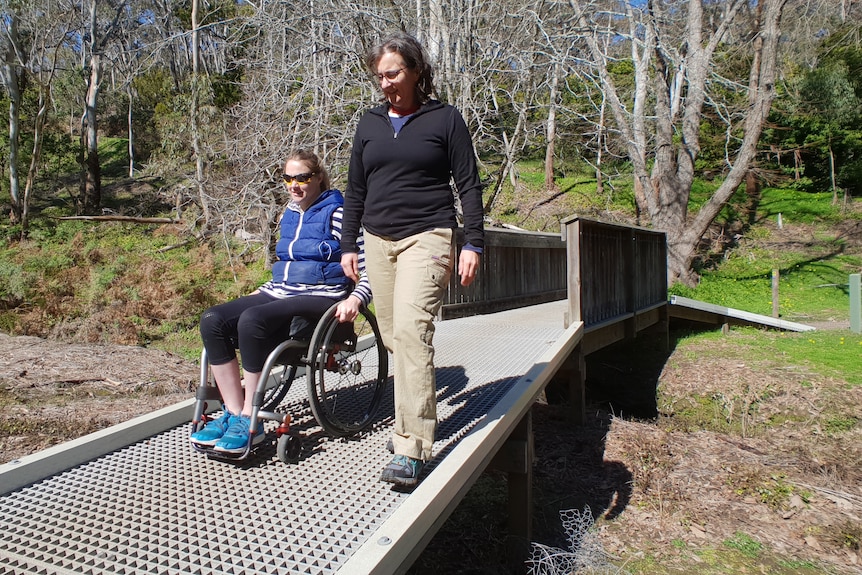 Yvette in a wheelchair, accompanied by a woman, both on bridge in middle of park or forest.