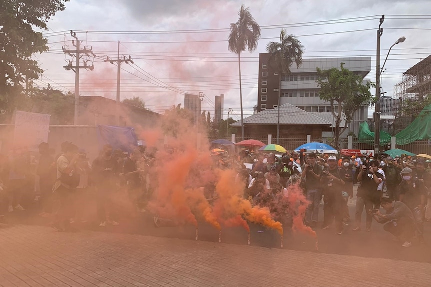 Thick orange smoke pours from flares on the ground in front of a crowd of people carrying colourful umbrellas.