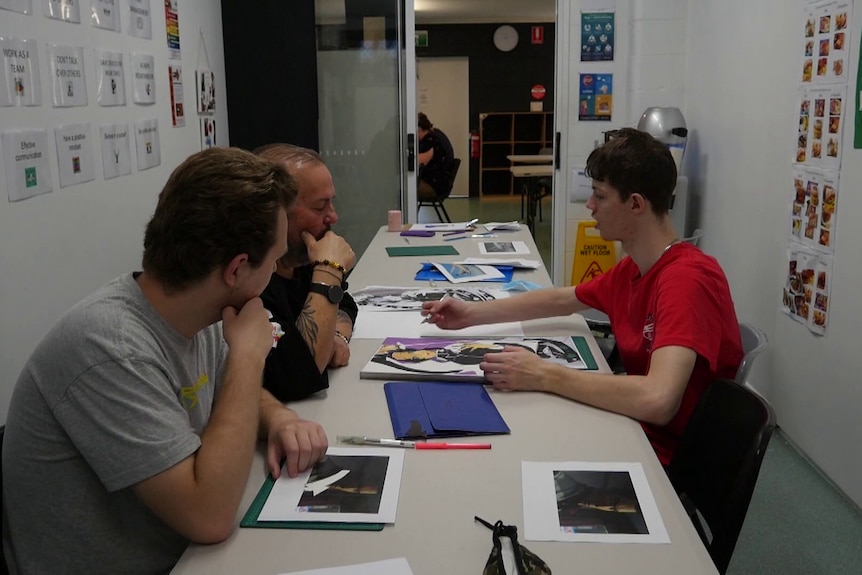 Three men sit down doing art at a table. 