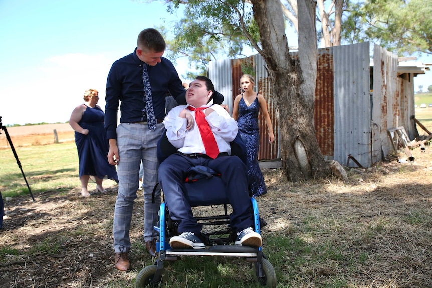 Two young men dresses in formal attire pose on a regional property. One man is wheelchair bound.