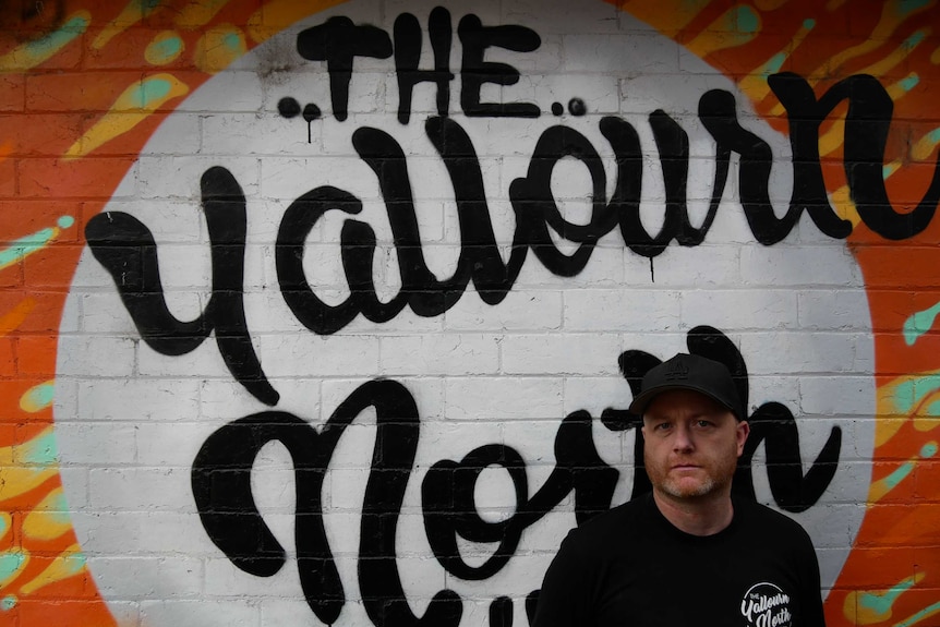 A man in a dark cap and shirt standing against a spray painted brick wall.