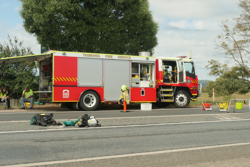 A fire truck with open compartments is parked beside a highway. Two people sit in chairs behind it