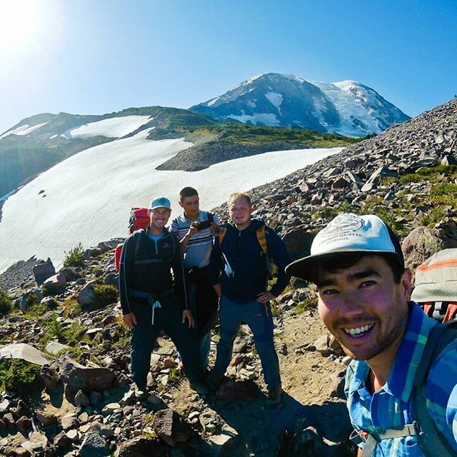 A man smiles as he takes a picture of himself with three friends atop a mountain.