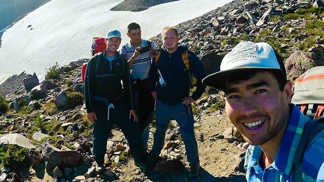 A man smiles as he takes a picture of himself with three friends atop a mountain.