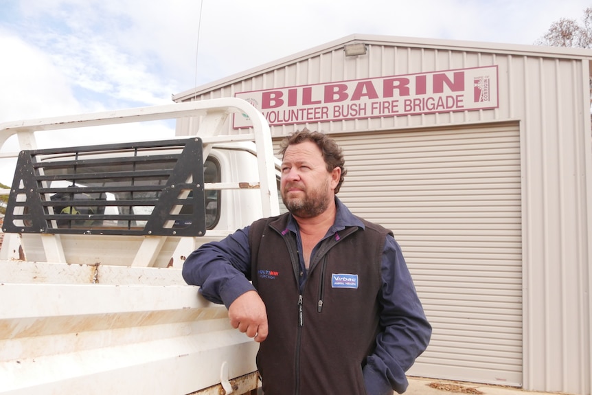 a man leans on a ute in front of a fire shed