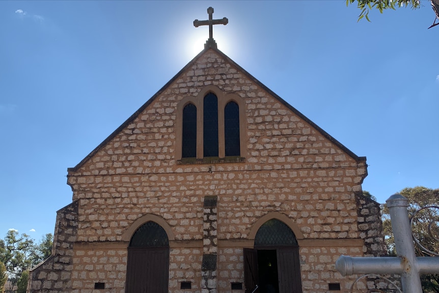 The sun shines behind a cross atop a sandstone building with two doors at the bottom and large cathedral windows 
