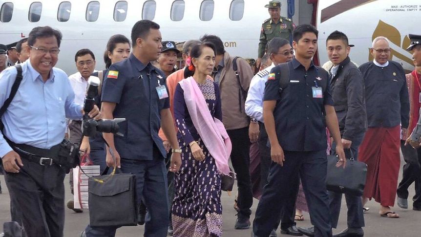 Aung San Suu Kyi is surrounded by military people and officials at an airport.