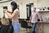 A female hairdresser wearing a face mask cuts a woman's hair, while a man wearing a face mask watches on.