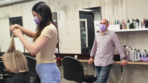 A female hairdresser wearing a face mask cuts a woman's hair, while a man wearing a face mask watches on.