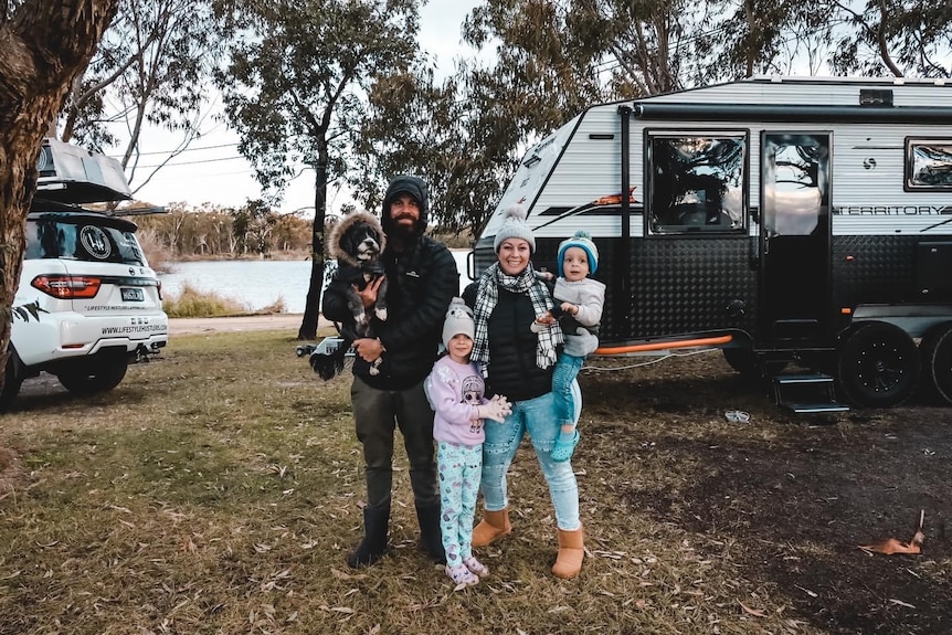 A mother, father, two children and their dog rugged up in warm clothes standing in front of a caravan.