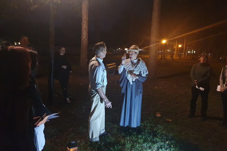 A man and woman in Victorian costume in front of trese at night while people watch. Woman wears hat, blue gown.