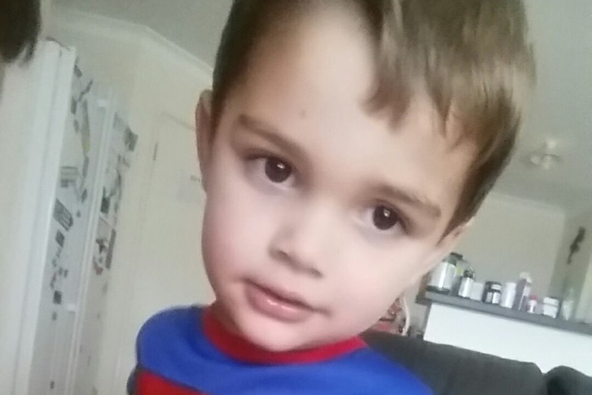 Two-year-old Lachlan Mitchell looks toward the camera wearing a Superman costume indoors.