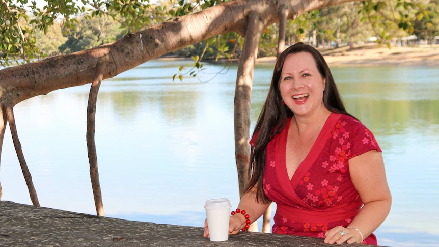 A woman with long, dark hair, wearing a red dress, sitting at a wooden bench under a tree near a river.