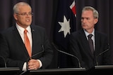 Three men stand behind podiums in front of Australian flags.