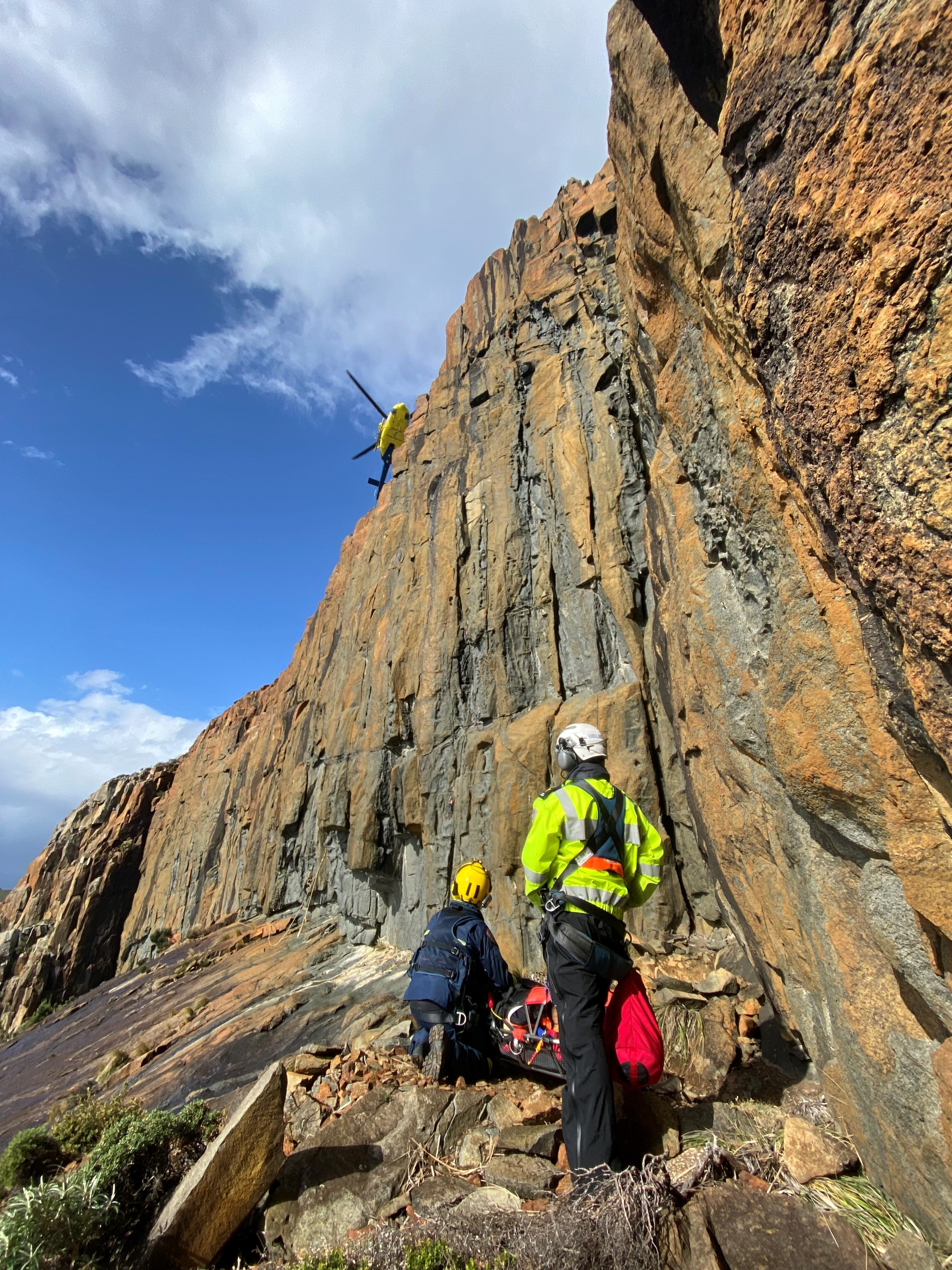 Two rescue paramedics or police officers wearing helmets and safety harnesses stand on a cliff face