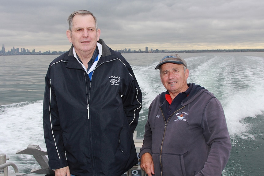 Bob Pearce and Tony Spinelli stand at the back of a boat on an overcast day. The Melbourne skyline is in the distance.