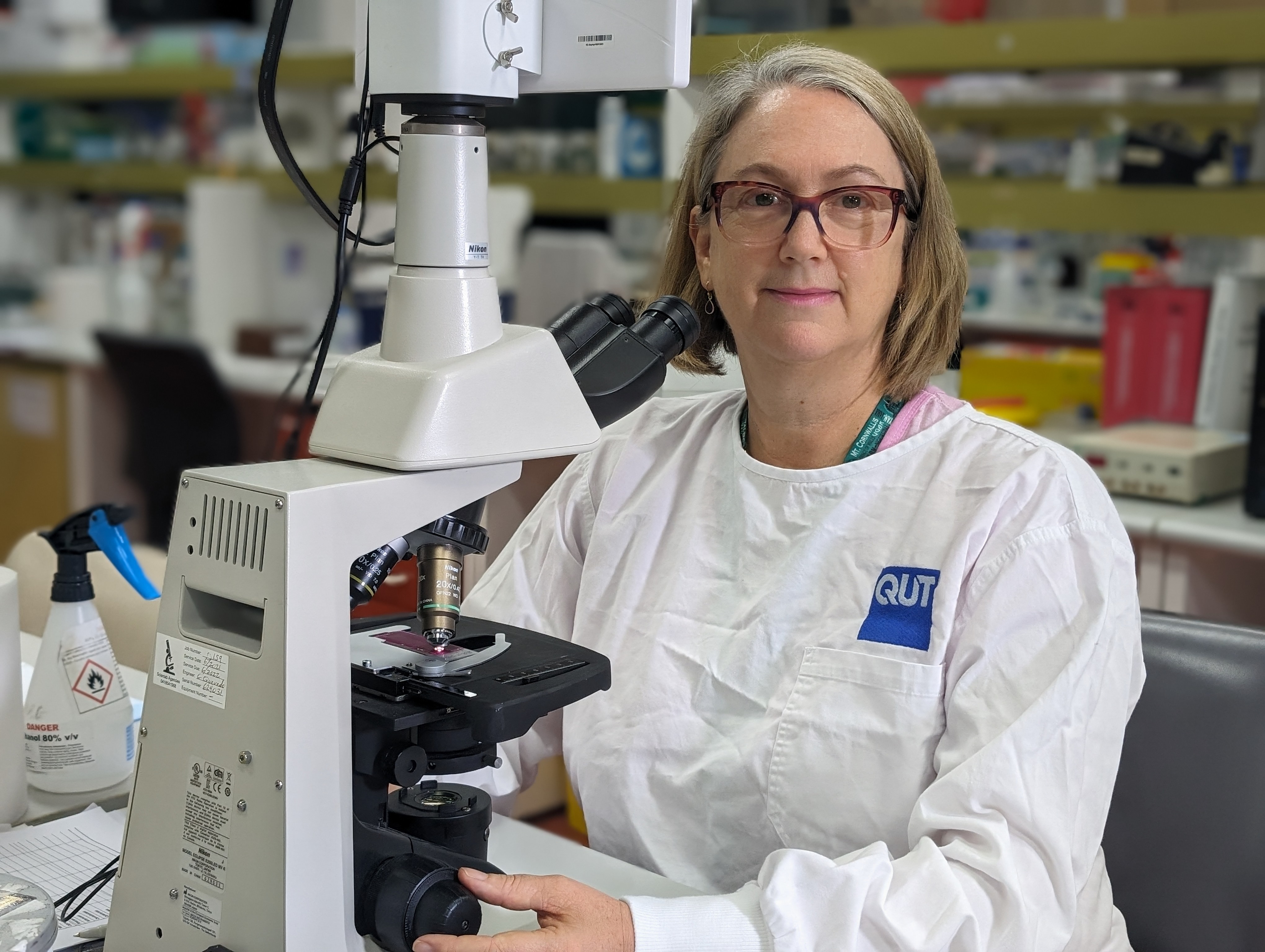A female, middle-aged white woman sitting in a laboratory. She is wearing a white lab coat