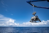 A large seabed mining machine being dangled over the ocean.  