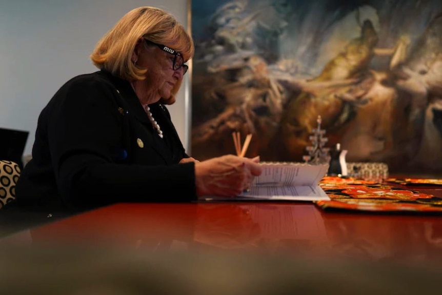 A woman sits at a desk in front of a painting.