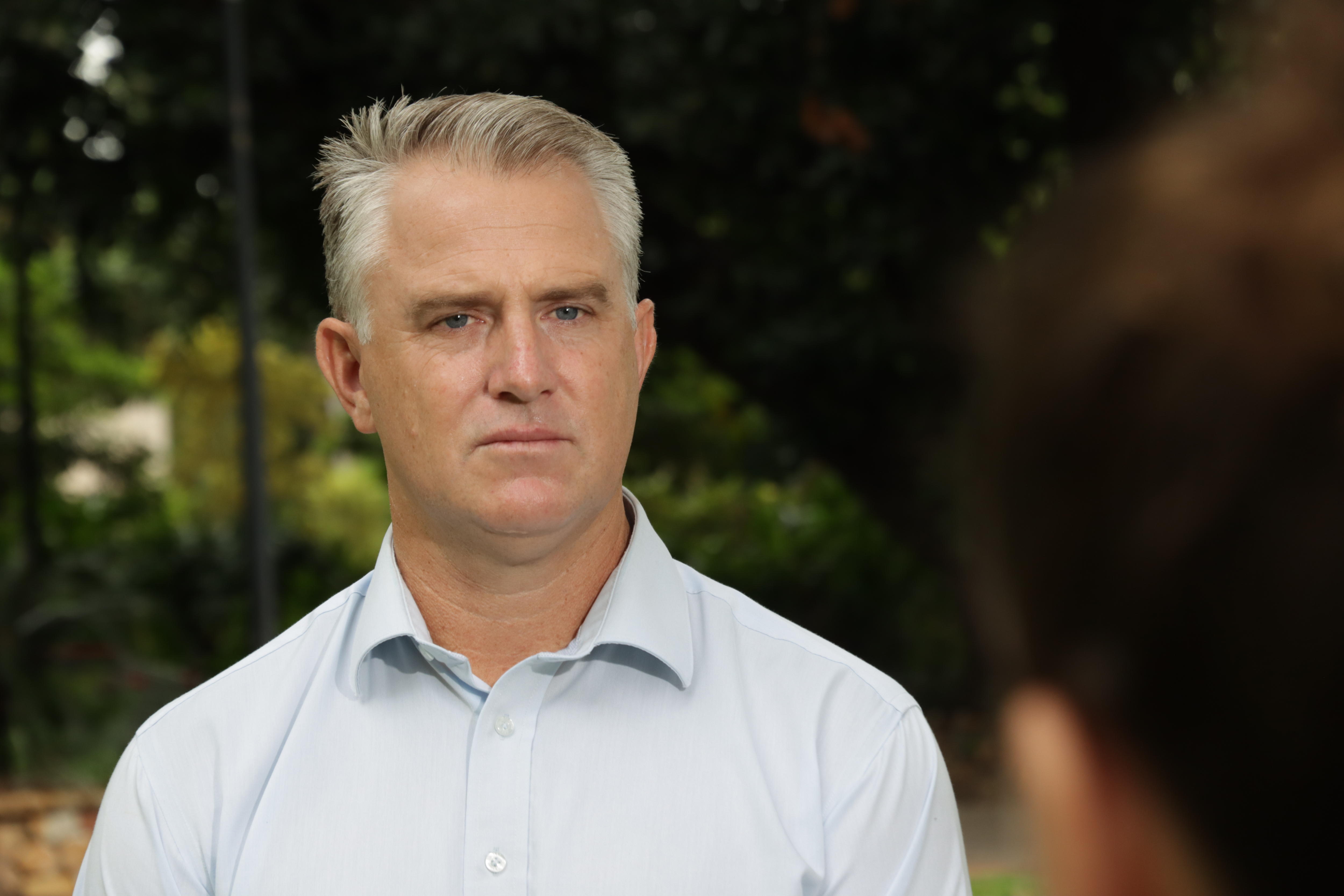  A man in a white shirt looks seriously at the camera. He is outdoors and talking to a reporter.