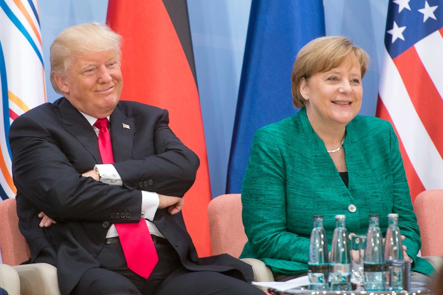 Donald Trump sits smiling with arms folded at a small table next to Angela Merkel.
