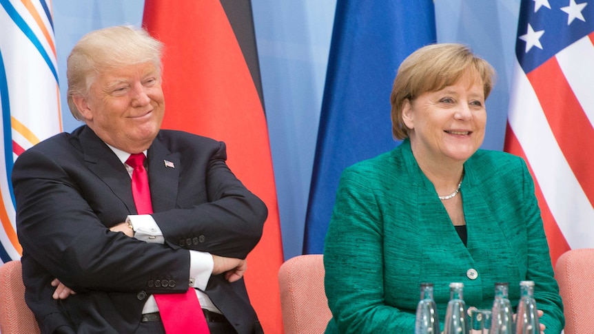 Donald Trump sits smiling with arms folded at a small table next to Angela Merkel.