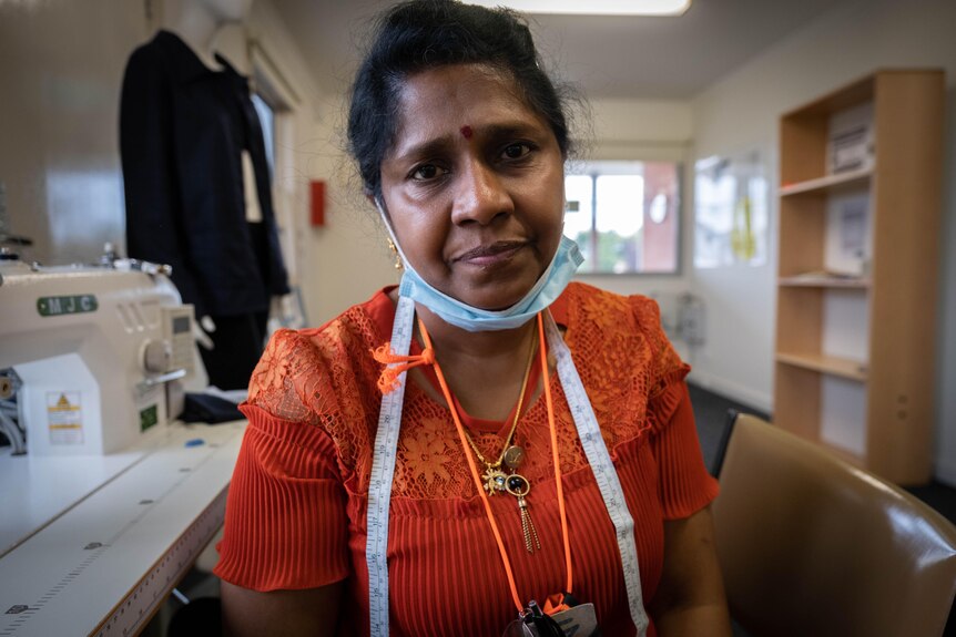 Rajani Nelson wears an orange top sits next to a sewing machine looking into the camera.
