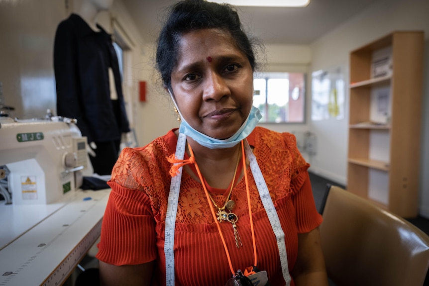 Rajani Nelson porte un haut orange est assis à côté d'une machine à coudre et regarde l'appareil photo.