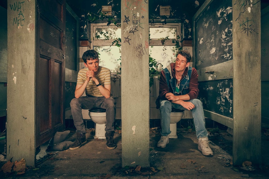 Two teenage boys sitting in rundown toilet stalls