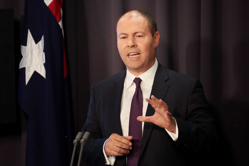 Balding middle aged man wearing a suit and tie speaking while standing at a lectern in front of a draped Australian flag