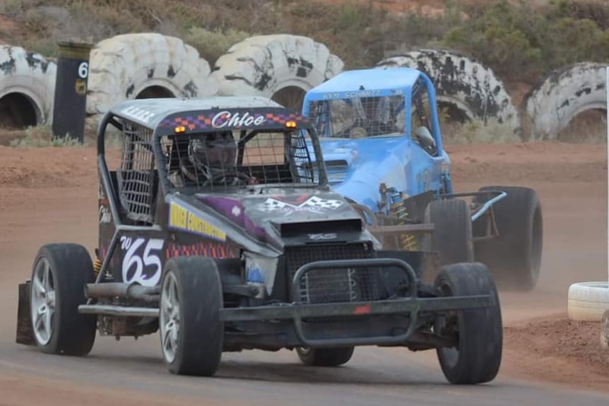 Black racing car in front of blue one on dirt track with big white tyres in background