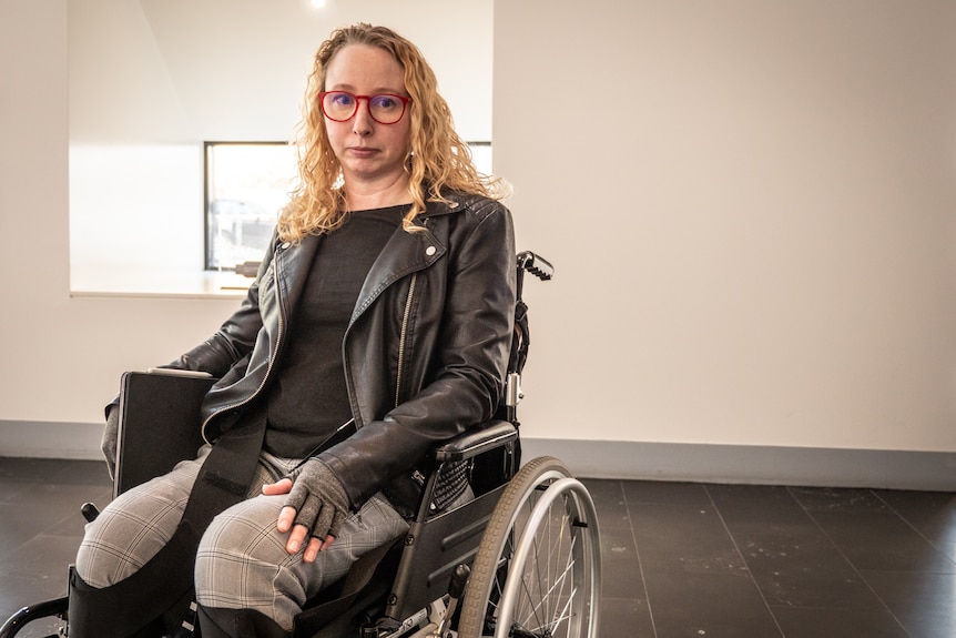 Woman with blonde hair and black jacket in wheelchair in front of white wall and window