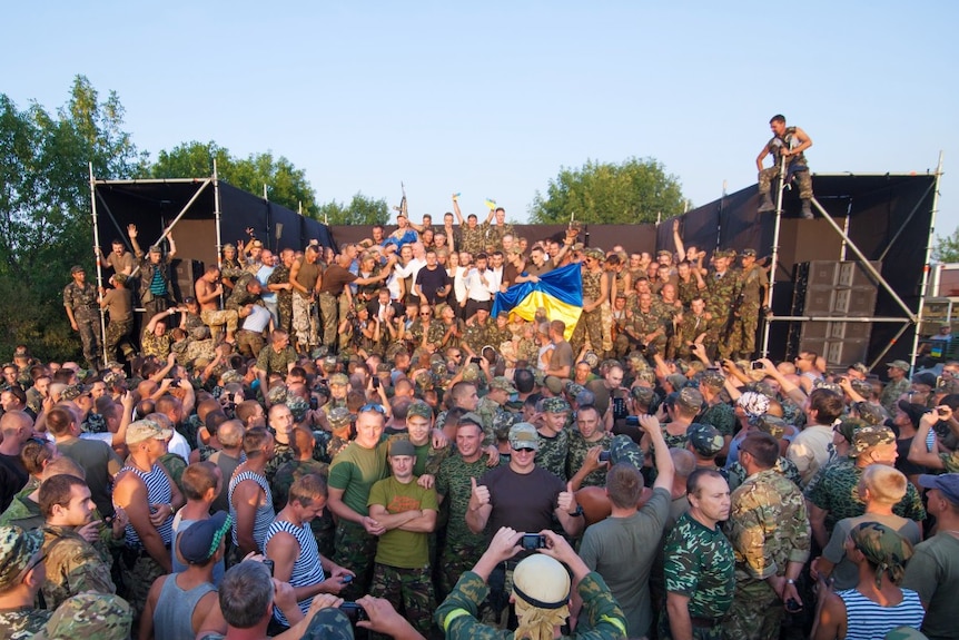 A large group of people on a stage, performing to a large crowd of soldiers, seen from a distance.