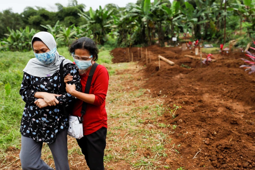 Two women wearing masks and looking down walk away from a grave site.