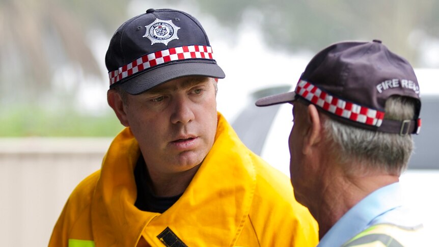 Two men stand side by side talking, both of them wear the same blue fire brigade cap.