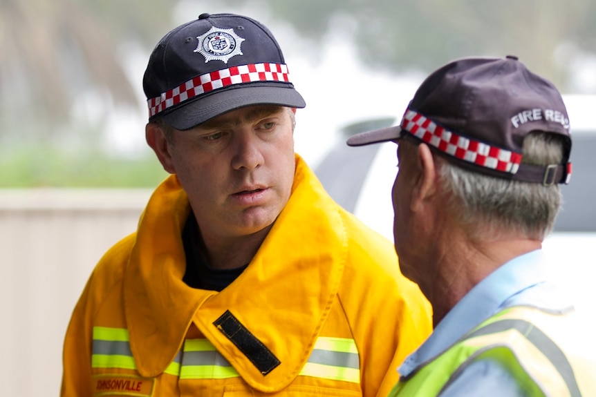 Two men stand side by side talking, both of them wear the same blue fire brigade cap.