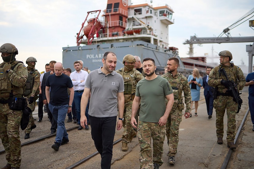 Men in army fatigues walk along a port with a ship in the background, flanked by armed soldiers.