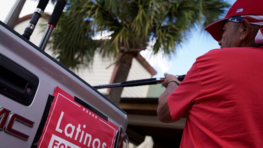 A man dressed in red puts something into the back of a car with a Latinos for Trump sign on the back.