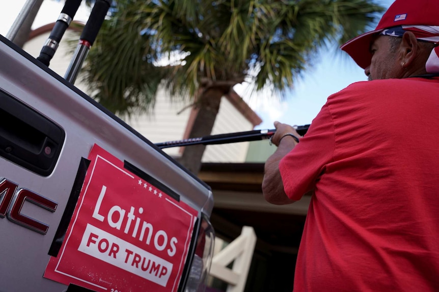 A man dressed in red puts something into the back of a car with a Latinos for Trump sign on the back.