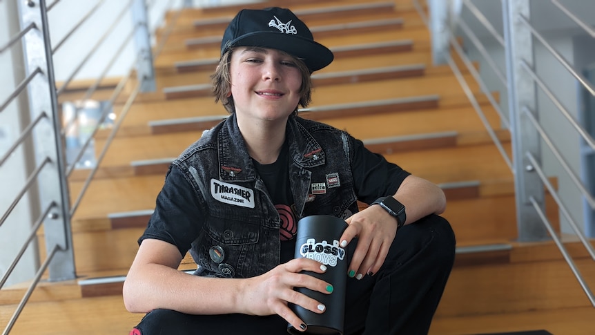 A boy sits on some steps holding a cylinder with the Glossy Boys label and showing his own polished nails