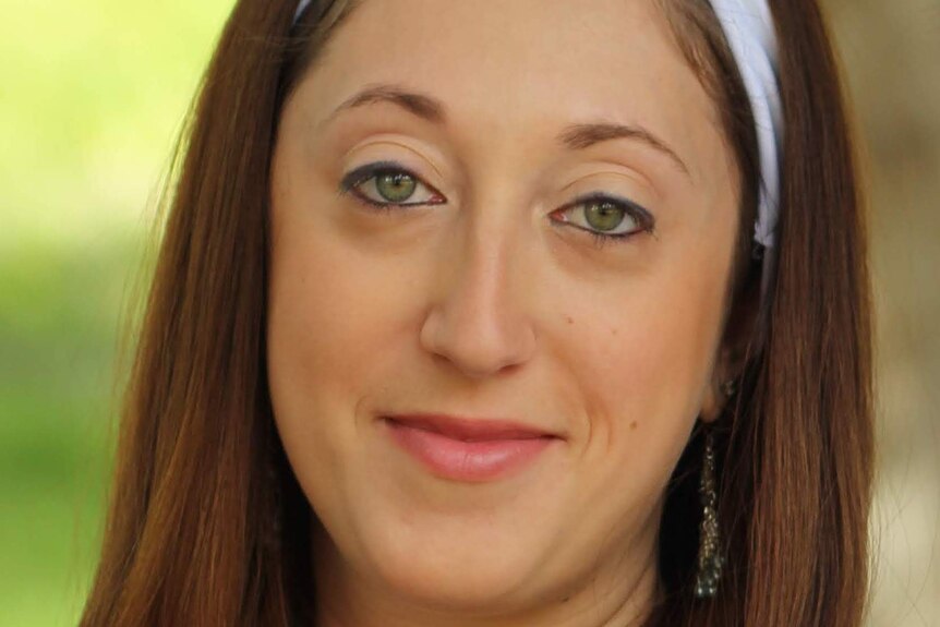 Shana Aaronson wears a white headband and smiles with closed lips as she stares at the camera