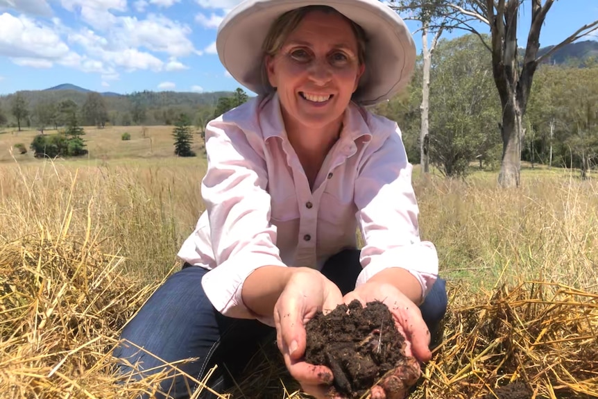 A lady crouches down holding out a handful of soil and worms towards the camera.